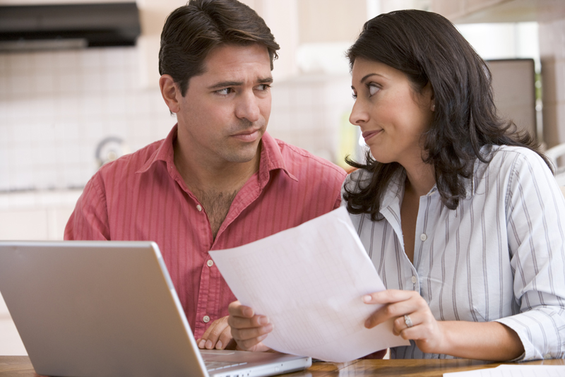 Couple in kitchen with paperwork using laptop looking unhappy